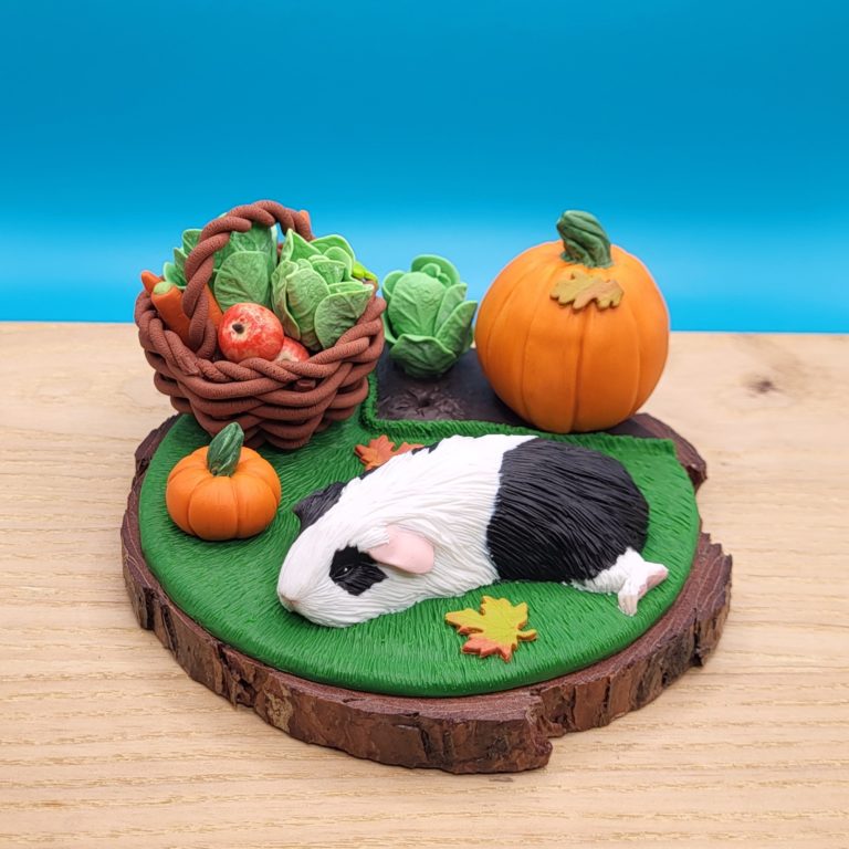 Black and white guinea pig sleeping on a grass next to a vegetable patch with a lettuce and large pumpkin. There's a brown basket with lettuce, carrots, apples already gathered. There's a small pumpkin between the basket and the piggy. Set on a wood slice base.