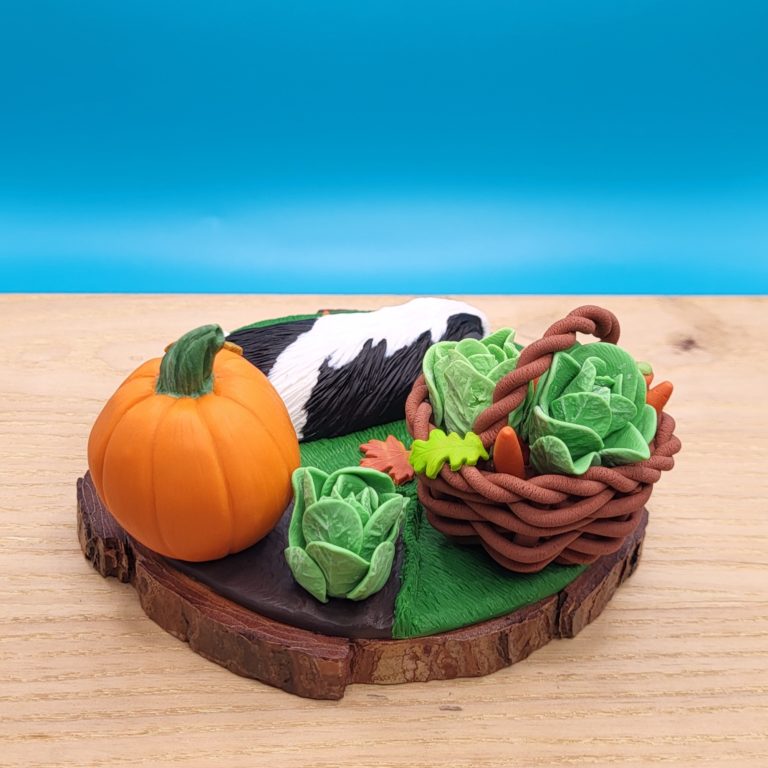 Black and white guinea pig sleeping on a grass next to a vegetable patch with a lettuce and large pumpkin. There's a brown basket with lettuce, carrots, apples already gathered. There's a small pumpkin between the basket and the piggy. Set on a wood slice base.