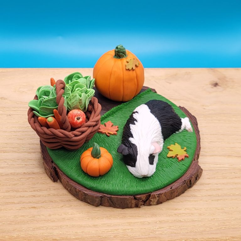 Black and white guinea pig sleeping on a grass next to a vegetable patch with a lettuce and large pumpkin. There's a brown basket with lettuce, carrots, apples already gathered. There's a small pumpkin between the basket and the piggy. Set on a wood slice base.