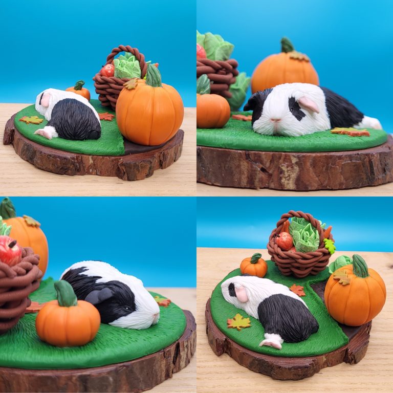 Black and white guinea pig sleeping on a grass next to a vegetable patch with a lettuce and large pumpkin. There's a brown basket with lettuce, carrots, apples already gathered. There's a small pumpkin between the basket and the piggy. Set on a wood slice base.