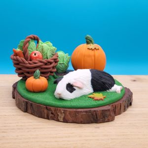 Black and white guinea pig sleeping on a grass next to a vegetable patch with a lettuce and large pumpkin. There's a brown basket with lettuce, carrots, apples already gathered. There's a small pumpkin between the basket and the piggy. Set on a wood slice base.