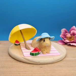 Long-haired cream guinea pig wearing a pastel blue hat with a white ribbon and pink bow, lounging on a stripy white and lavender blanket. A white and yellow umbrella and three slices of watermelon adorn the scene. Guinea pig is set on a wooden base.