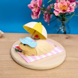 Long-haired cream guinea pig wearing a pastel blue hat with a white ribbon and pink bow, lounging on a stripy white and lavender blanket. A white and yellow umbrella and three slices of watermelon adorn the scene. Guinea pig is set on a wooden base.