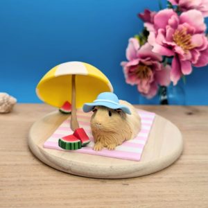 Long-haired cream guinea pig wearing a pastel blue hat with a white ribbon and pink bow, lounging on a stripy white and lavender blanket. A white and yellow umbrella and three slices of watermelon adorn the scene. Guinea pig is set on a wooden base.