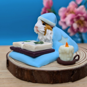 Clay figurine of a guinea pig wearing a blue nightcap and matching blanket with star patterns, sitting on a round wooden base. The guinea pig is looking at an open book with illustrations of lettuce and kale. Beside it, there is a lit candle in a small brown holder