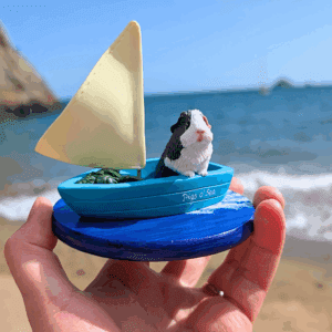 Guinea Pig Sculpture featuring white and grey piggy in a sailing boat looking out into the distance. There's a crate of spinach leaves for snacking.