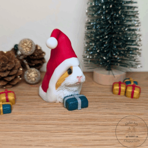 A handmade Christmas Gnome Guinea Pig sculpture crafted from coloured polymer clay. The guinea pig is sitting upright, wearing a red Santa-style hat with a white trim and pom-pom, and holding a small wrapped present.
